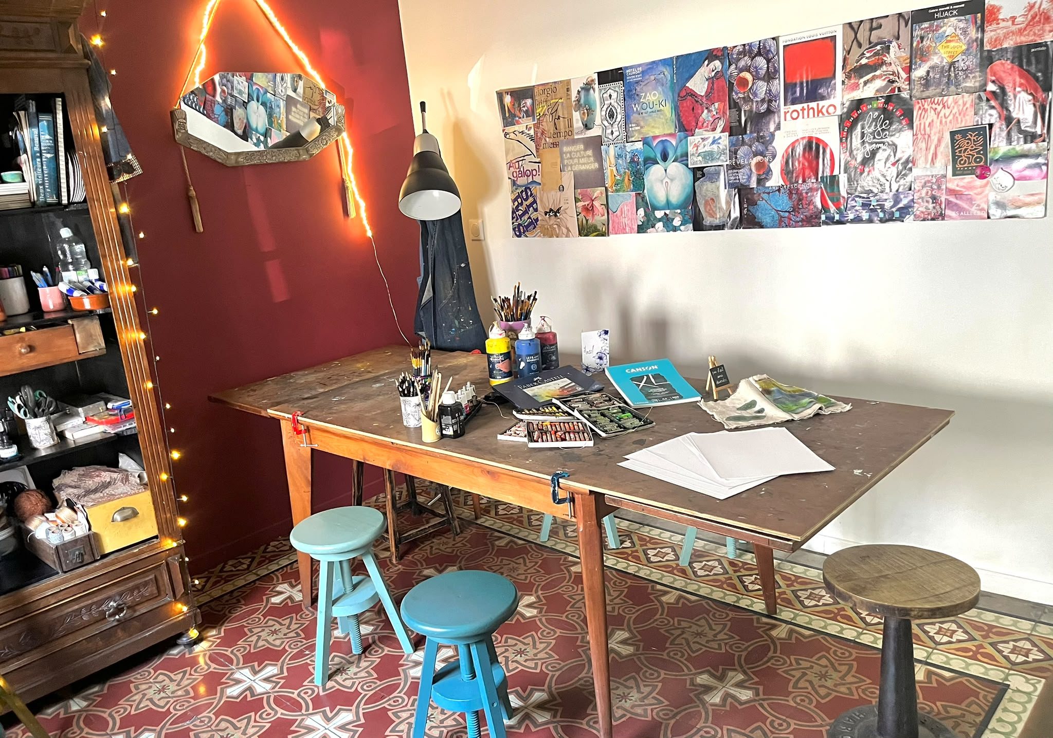La salle de l'atelier : grande table de bois patinée entourée de tabourets, armoire ancienne remplie de pinceaux et de peintures, mur bordeaux et carreaux de ciment