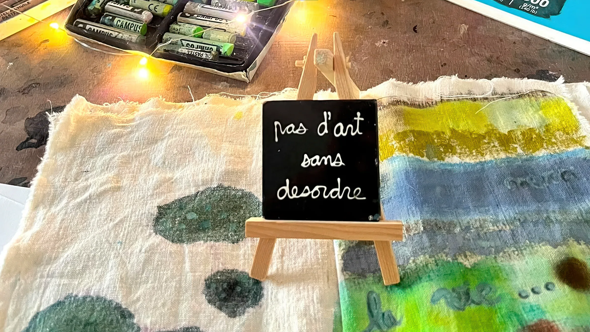 Une ardoise posée sur un petit chevalet de bois, où est écrit à la craie « pas d'art sans désordre », sur une toile peinte de bleus, de verts et de jaunes