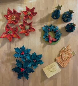 Ensemble de pieces en argile emaillee posees sur une table de l atelier Maison Maria Stella : fleurs de poinsettia rouges, branches de houx bleues, couronne, petits sapins et une colombe.