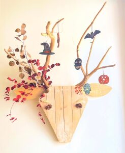 Sculpture murale figurant une tete de cerf assemblee en planches de bois brut, les bois faits de branches et decores de feuillages rouges, d une chauve-souris et d une citrouille.